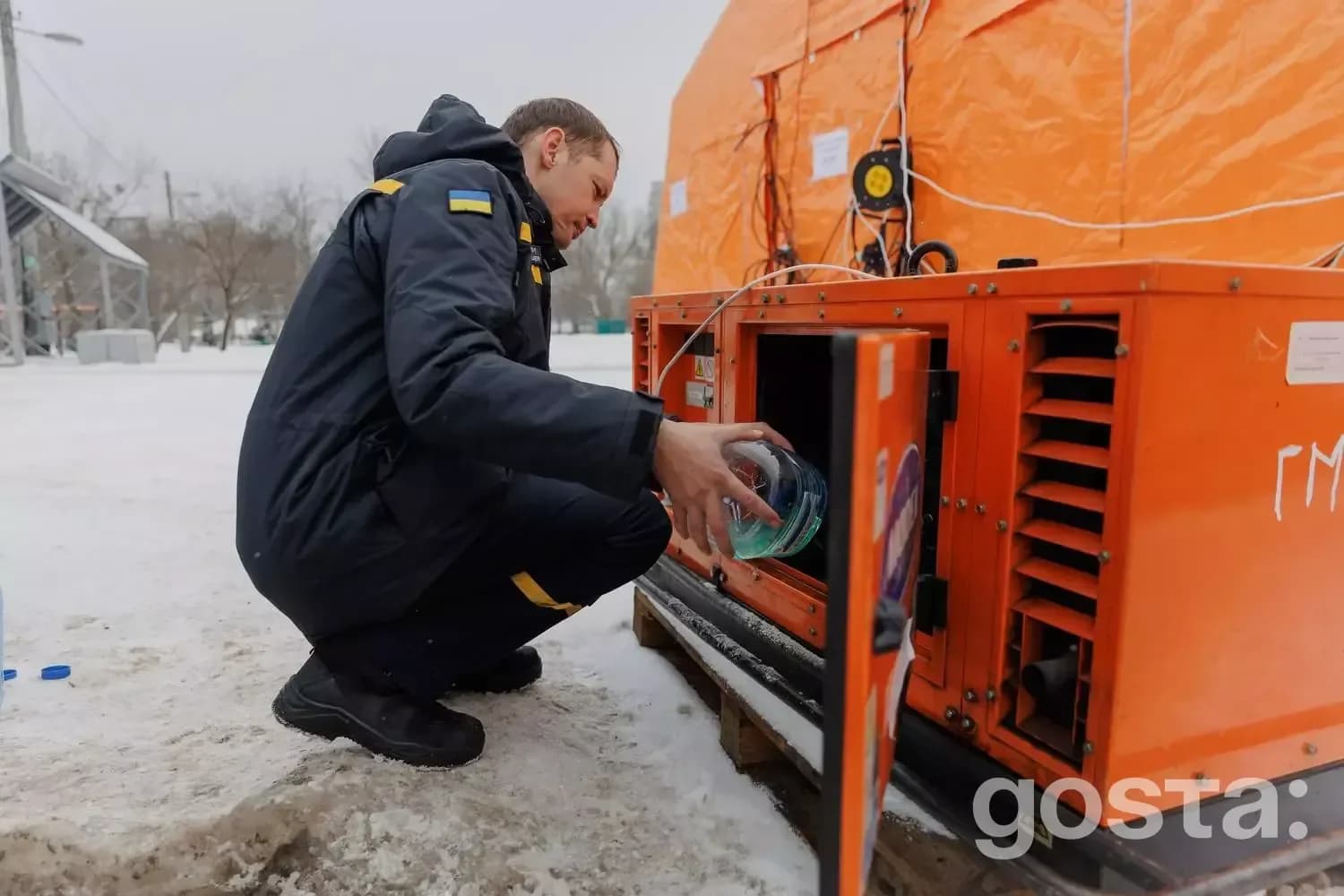 Людина в зимовому одязі активно працює біля генератора на снігу поруч з великим наметом.