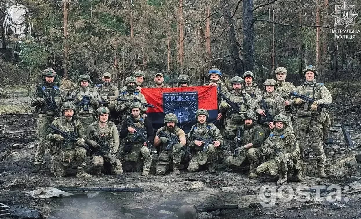 На зображенні група військовослужбовців стоїть в лісі з прапором.