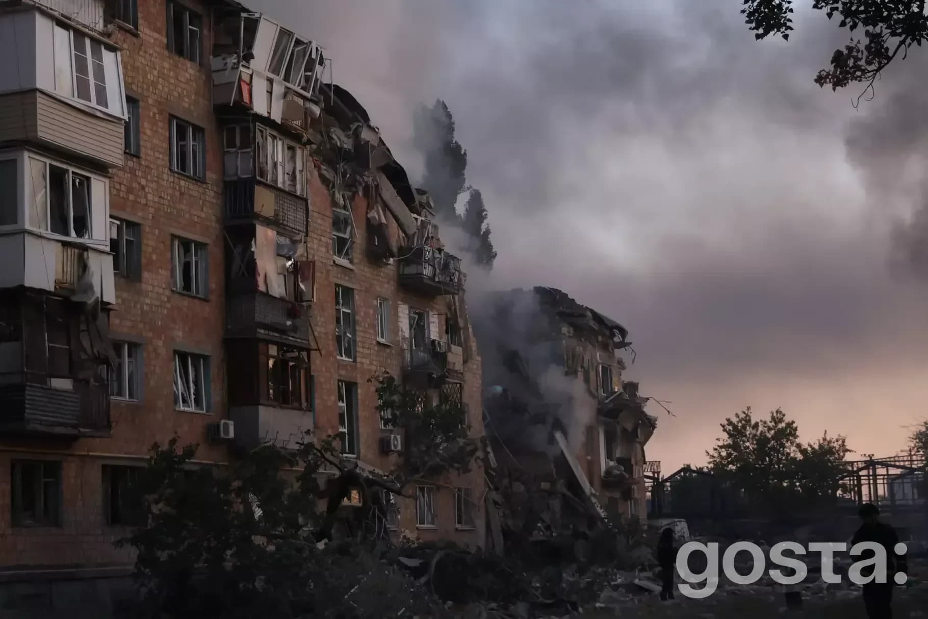 Зруйнована будівля після обстрілів в Україні, дим піднімається в небо, зруйновані фасади, в сутінковий час.