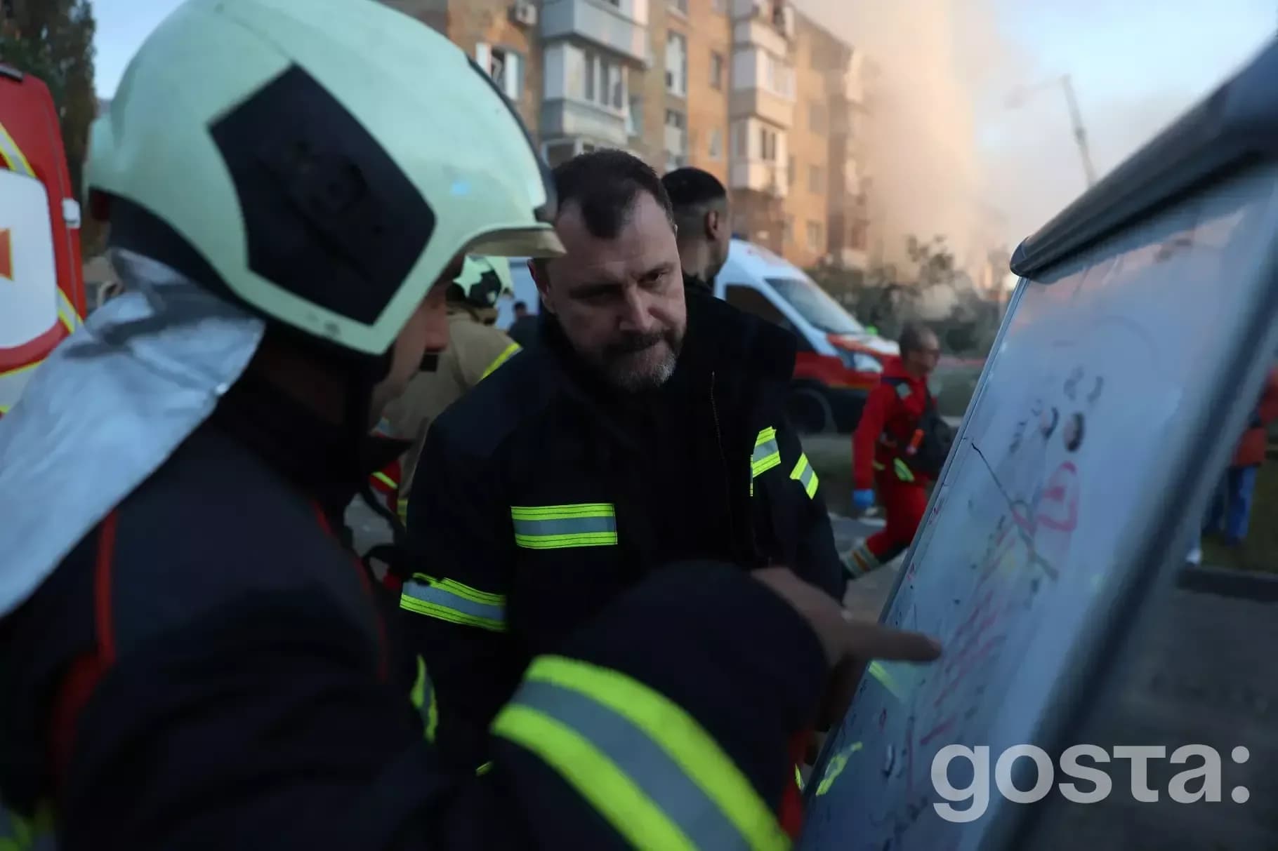 На зображенні пожежники обговорюють план дій біля багатоповерхового будинку з димом на тлі, Україна.
