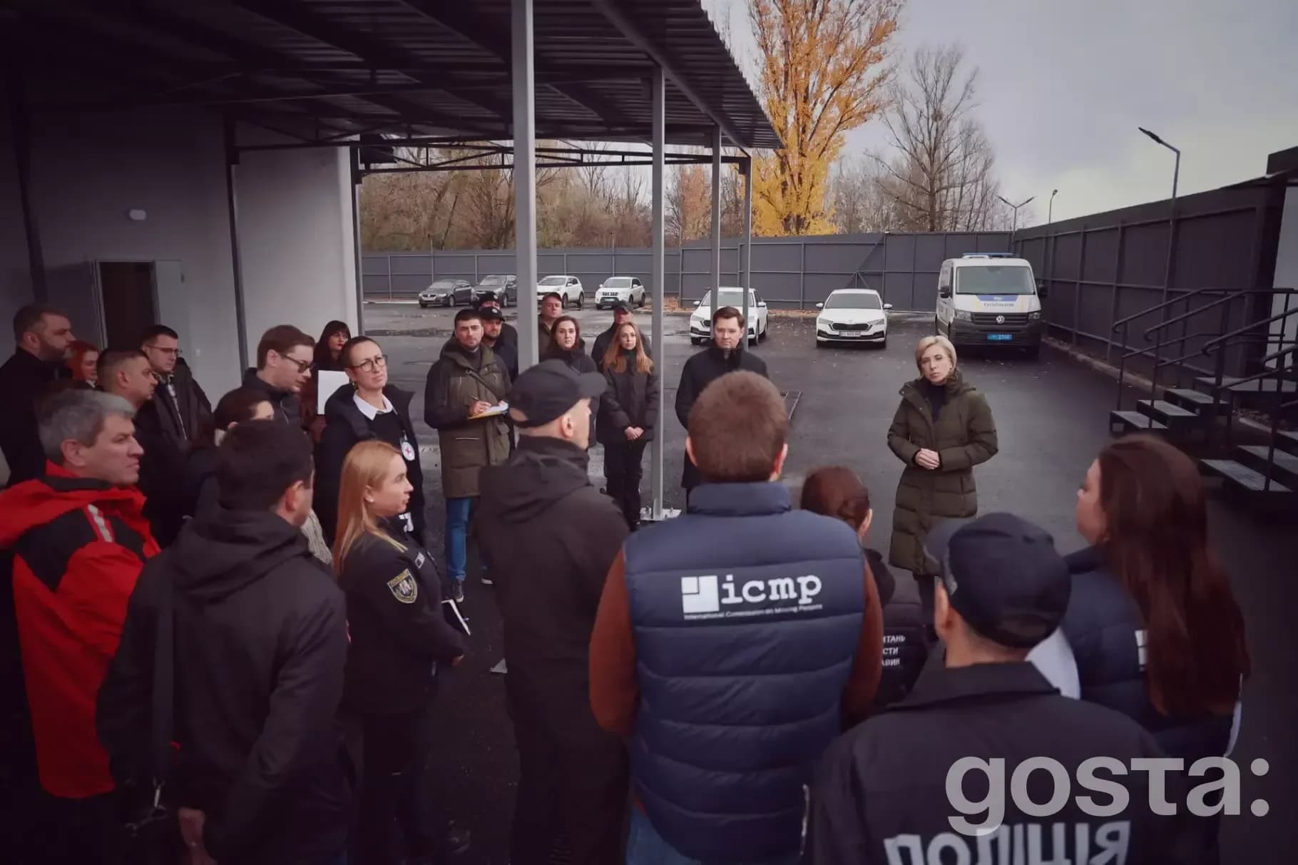 На зображенні люди в поліційній уніформі на зустрічі на території з автомобілями й будівлею.