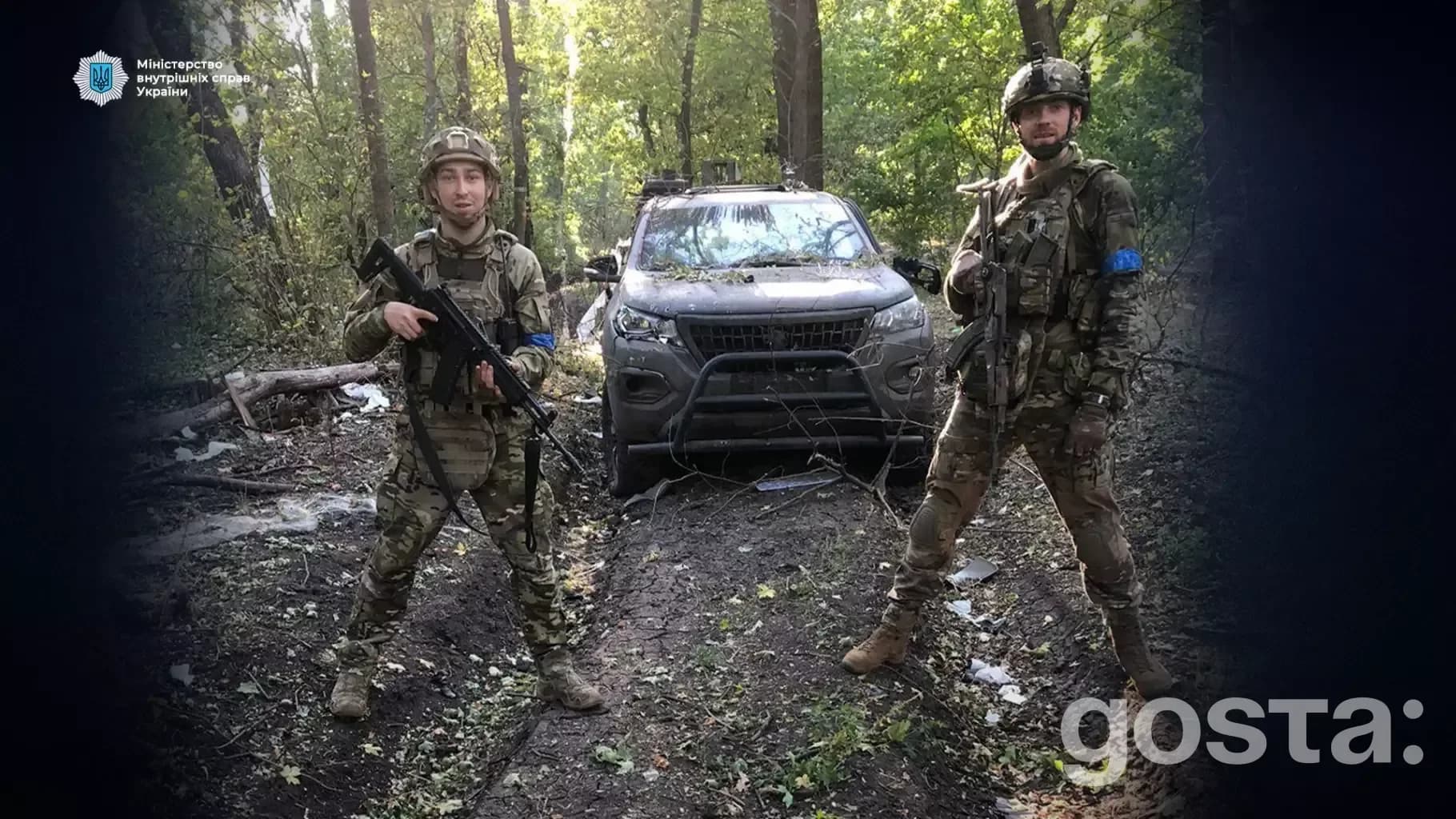 Двоє українських військових стоять у лісі перед позашляховиком, тримаючи зброю, у військовій уніформі.