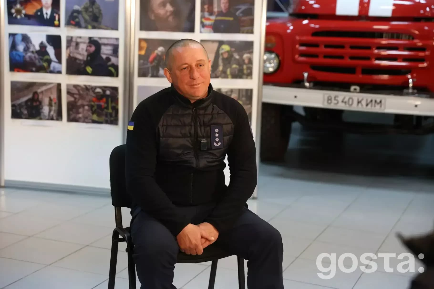 На фото рятувальник ДСНС України сидить на стільці у приміщенні, позаду стенд з фотографіями та пожежна машина.