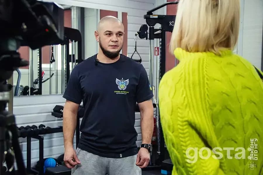 Чоловік у футболці Національної гвардії спілкується з жінкою в реабілітаційному центрі, тренажери на фоні.
