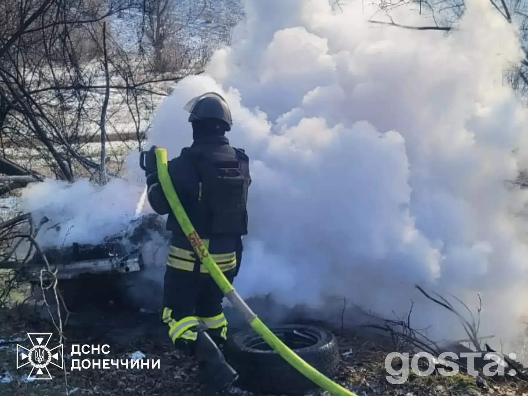 Пожежник гасить пожежу після обстрілів у Донецькій області, дим піднімається вгору, пошкоджена машина під деревами.