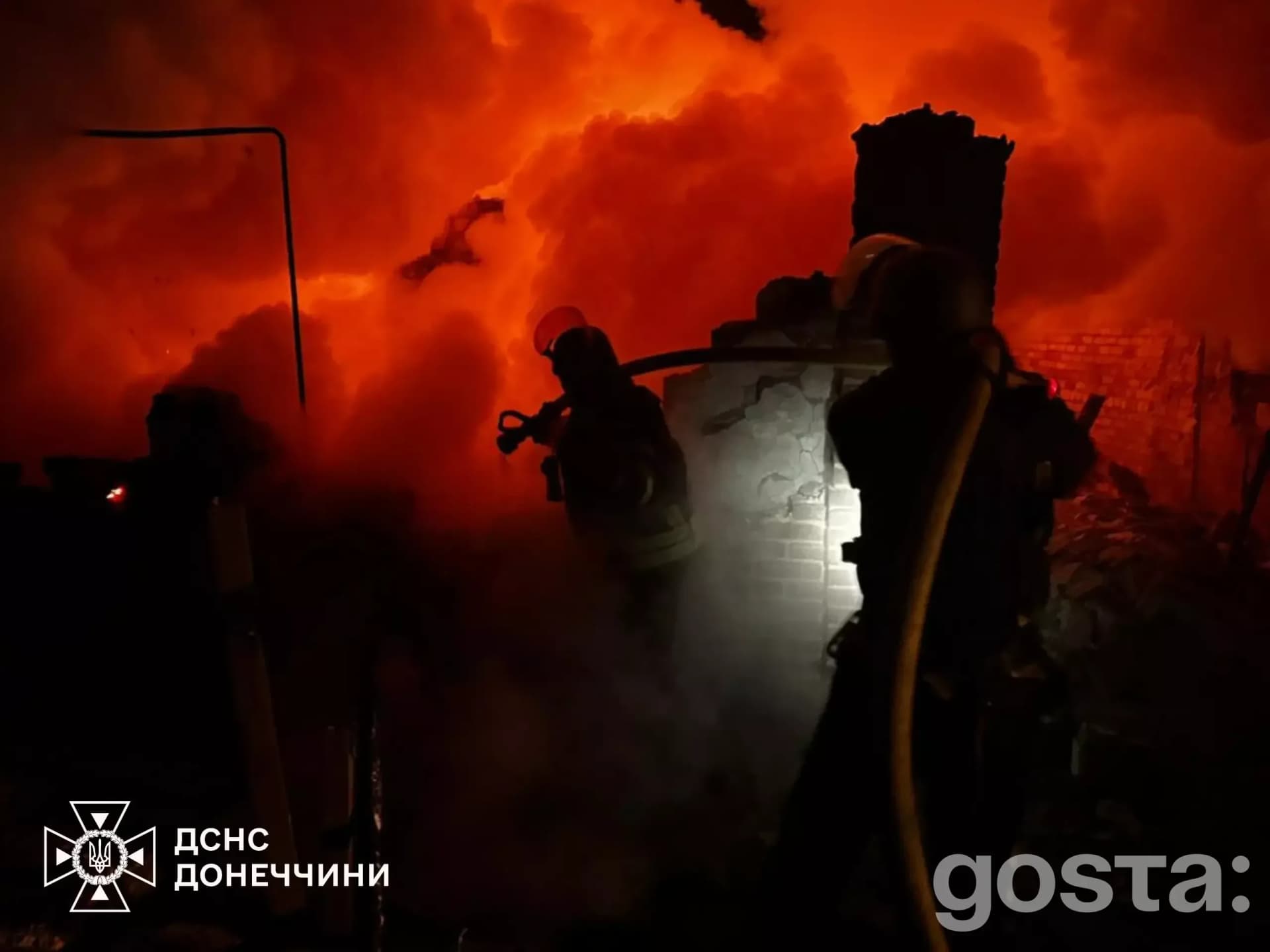 Рятувальники гасять палаючу будівлю на тлі яскравого полум’я після обстрілу на Донеччині.