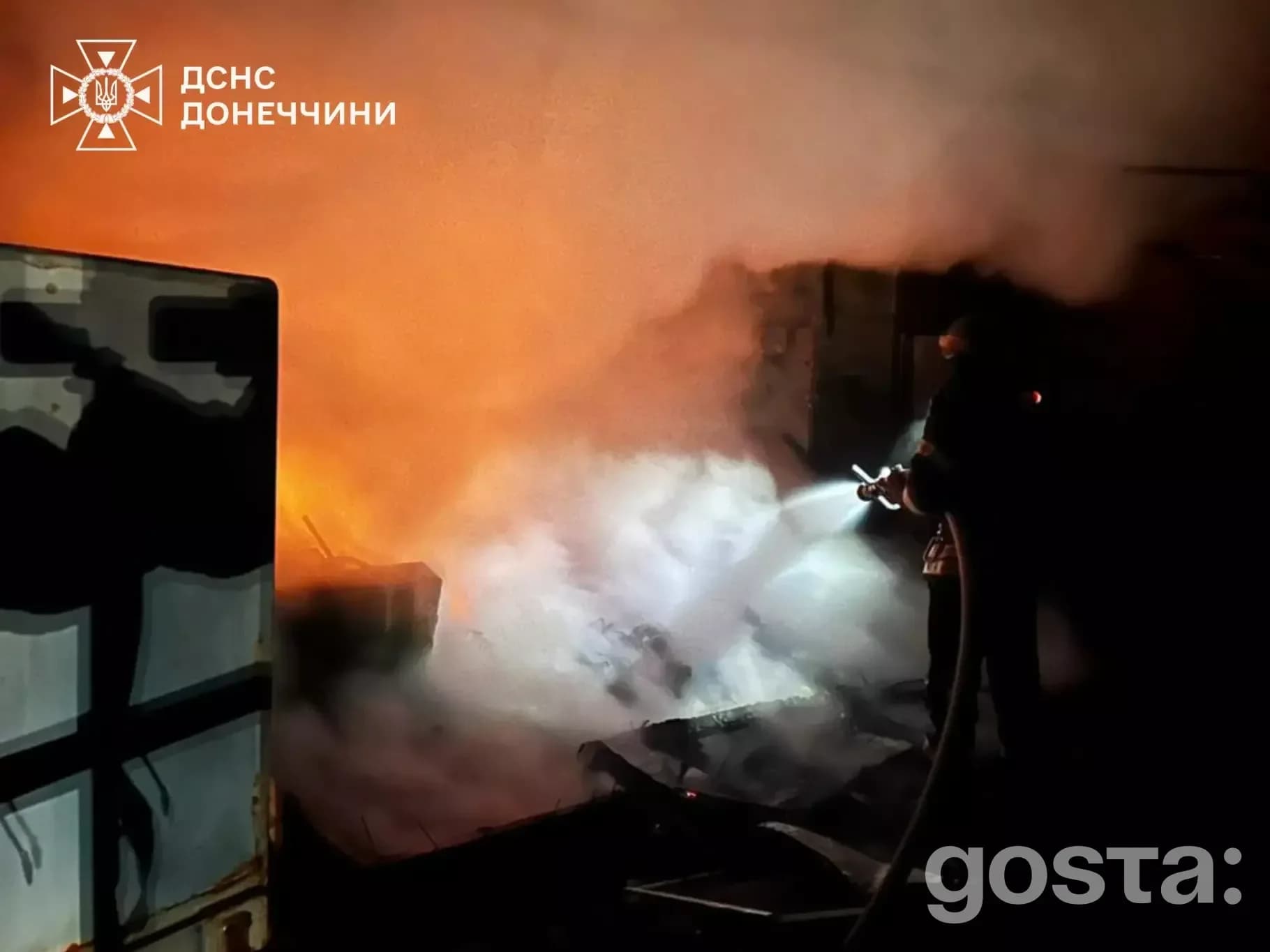 Рятувальник гасить пожежу після обстрілів на Донеччині, густий дим підсвічений полум’ям, нічна сцена з руйнуваннями.