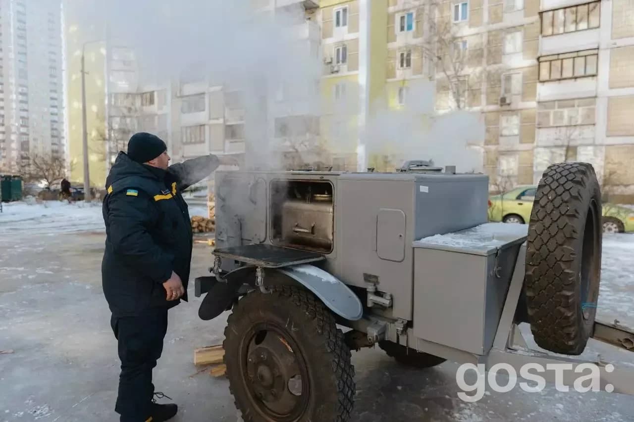 На зображенні рятувальник обслуговує мобільну пічку для обігріву на тлі багатоповерхівок.