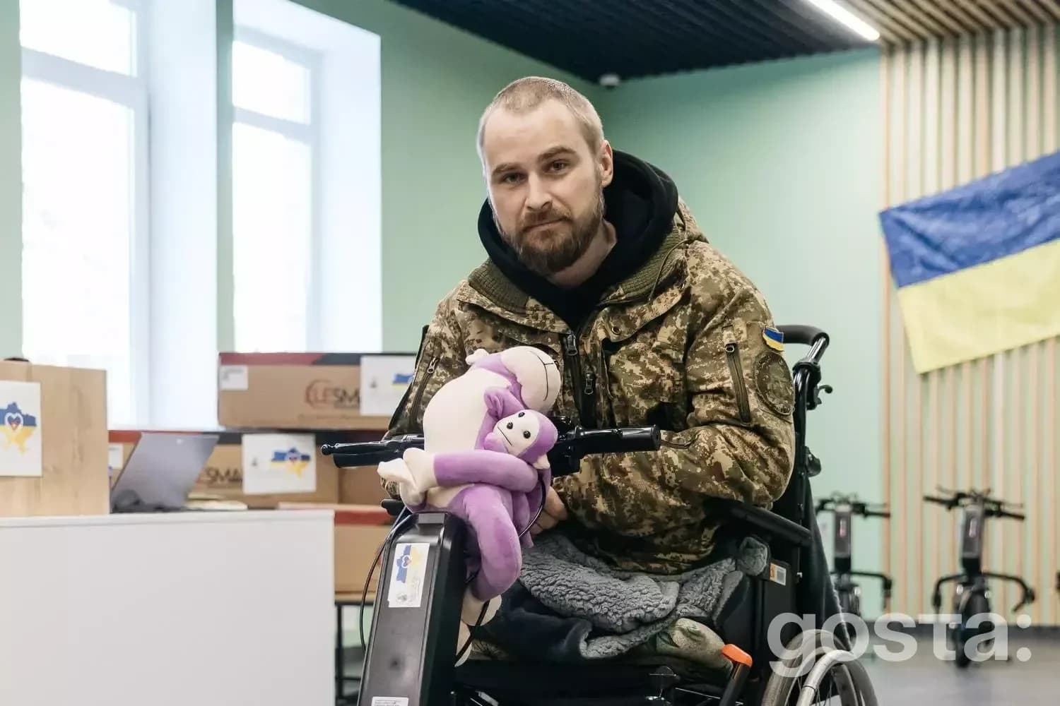 На зображенні: ветеран у камуфляжі сидить у візку з електроприводом, тримаючи м'яку іграшку, у приміщенні медичного закл