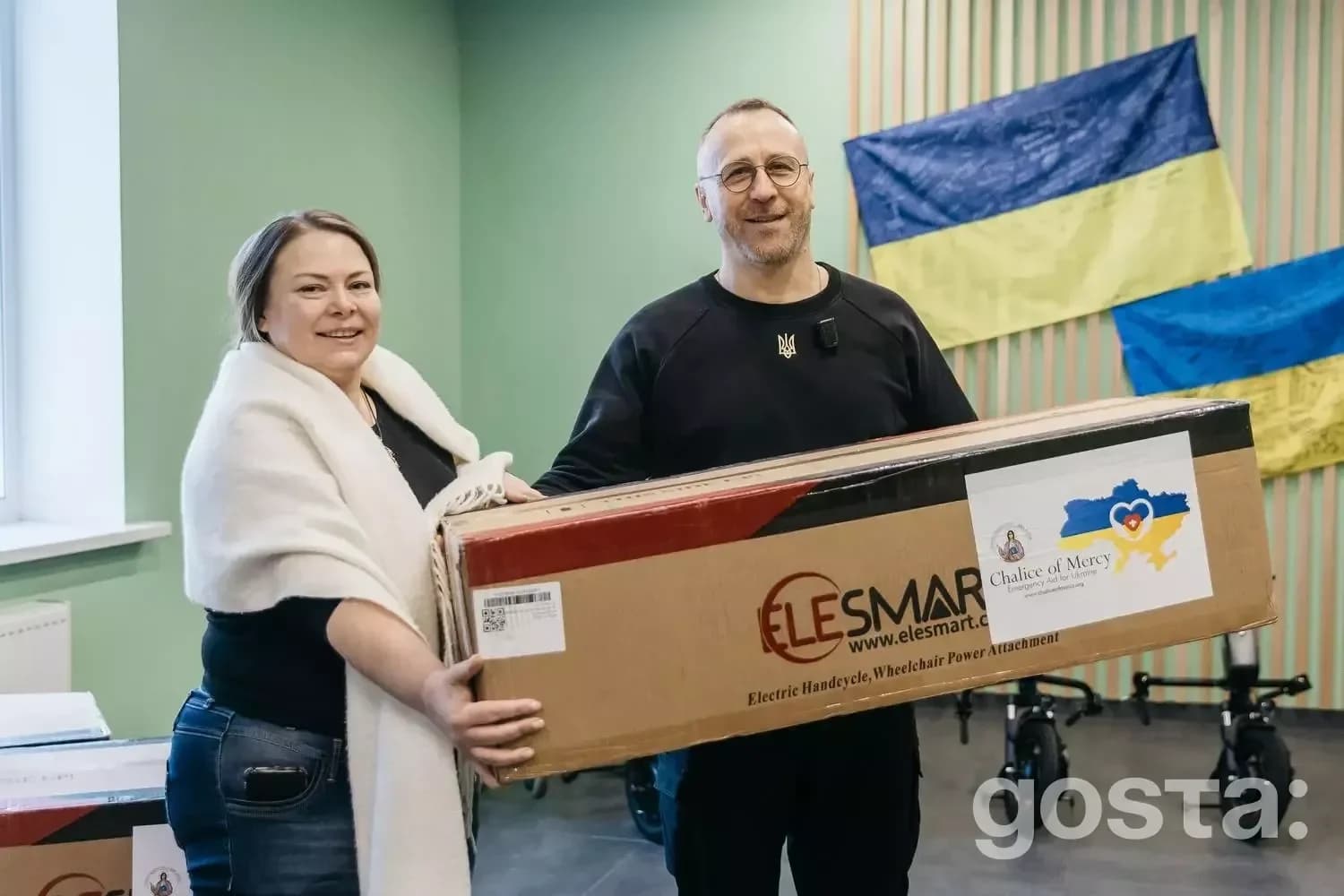 Я не можу впізнати людей на фото, але можу описати його. 

Двоє людей тримають коробку з електроприводом для крісла у це