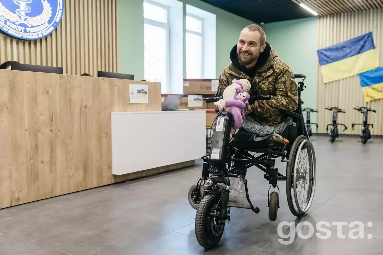 Людина в лікарні їде на електроколясці, посміхаючись, з плюшевою іграшкою на колінах.