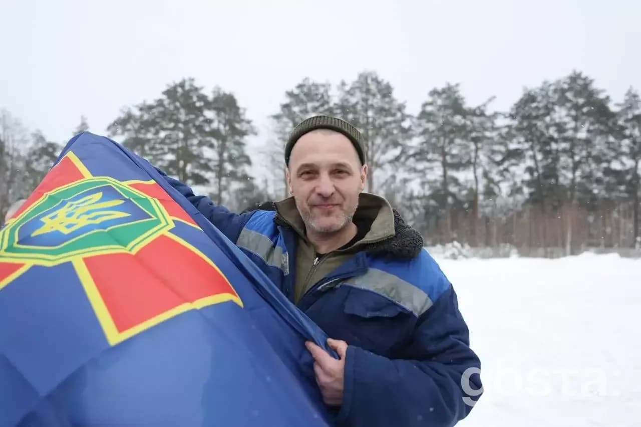 На фото чоловік у зимовому одязі з прапором України на фоні засніжених дерев.