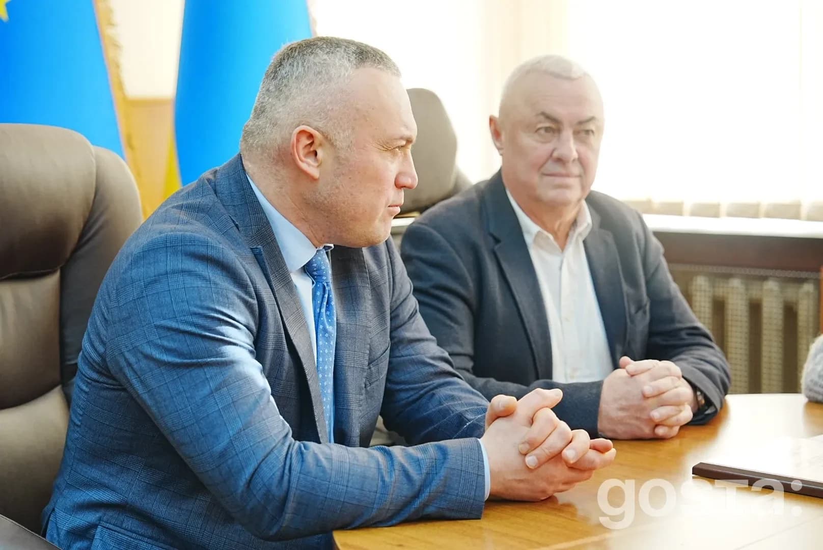 На зображенні двоє чоловіків у ділових костюмах сидять за столом під час офіційної зустрічі.