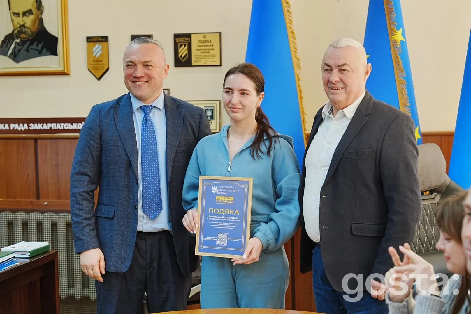 На фото: жінка в спортивній формі тримає почесну грамоту, поруч з нею двоє чоловіків у ділових костюмах.
