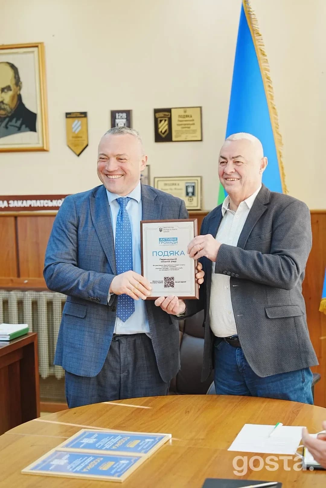 Двоє чоловіків під час вручення нагороди в кабінеті з українськими прапорами і грамотами на стіні.