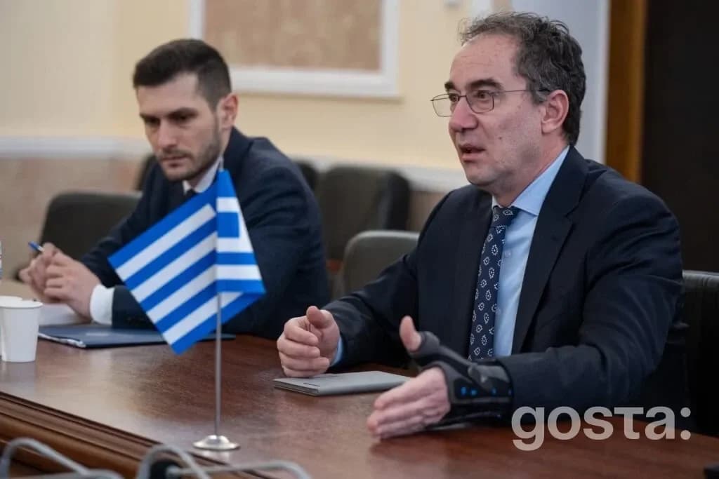 На зображенні двоє чоловіків за столом під час зустрічі з грецьким прапором, обговорюють культурну співпрацю.