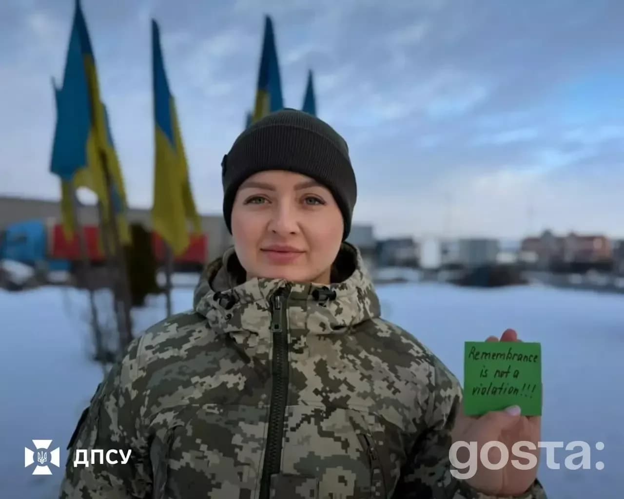 Українська військова у формі тримає напис "Remembrance is not a violation" на тлі прапорів України.