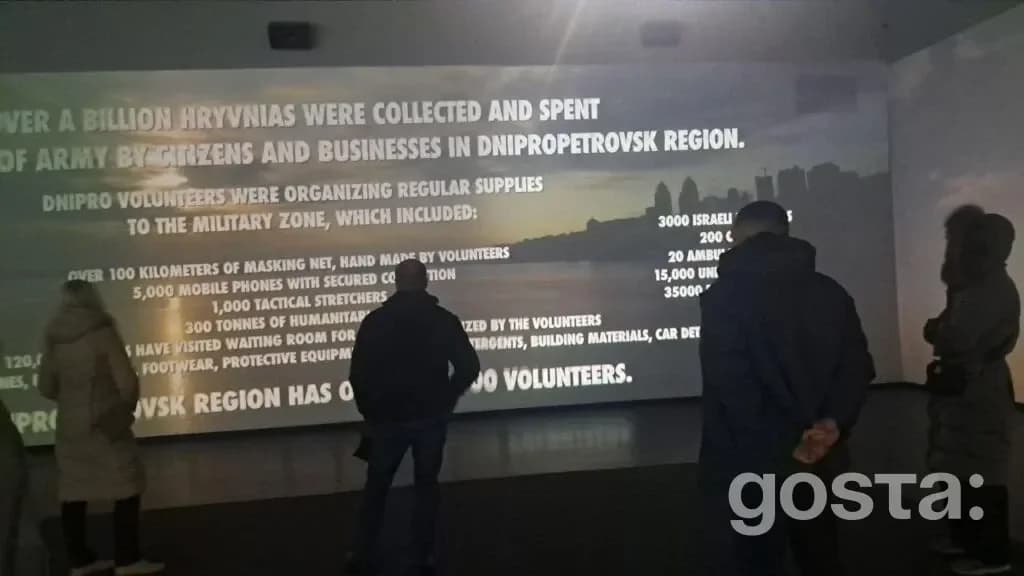 На зображенні відвідувачі переглядають освітлений інформаційний екран у SHELTER-ART SPACE, підземному просторі театру.
